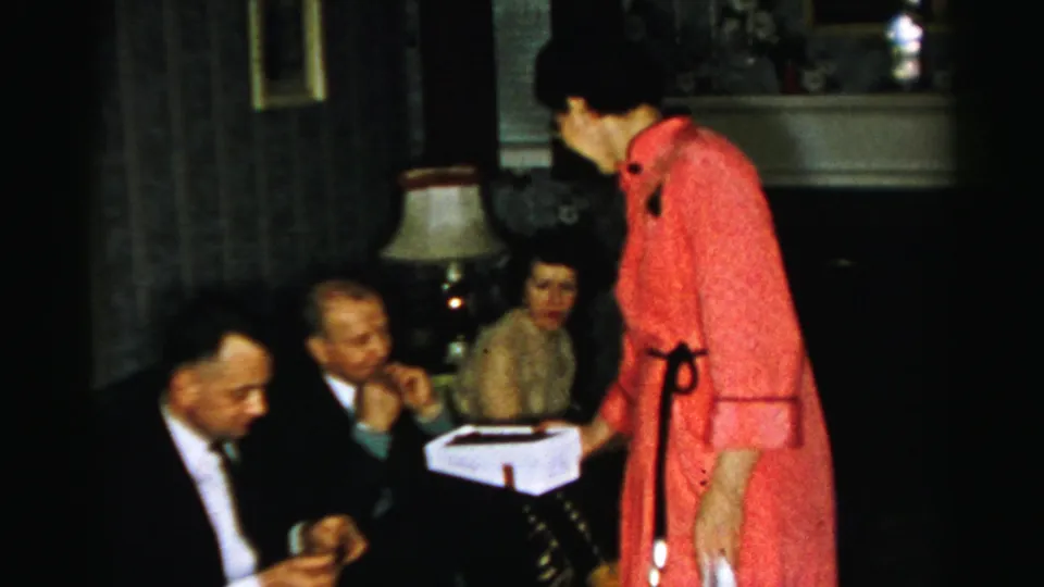 At the Albany indoor gathering, a woman in a red dress stands with a white box while seated adults watch or converse nearby.
