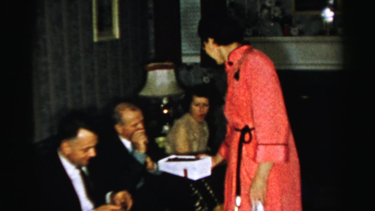 Albany-area 1950s home-movie frame showing a woman in a red dress offering a white box to suited men seated in a living room.