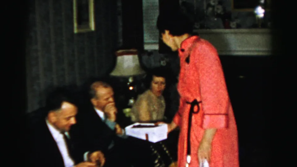 Albany-area 1950s home-movie frame showing a woman in a red dress offering a white box to suited men seated in a living room.