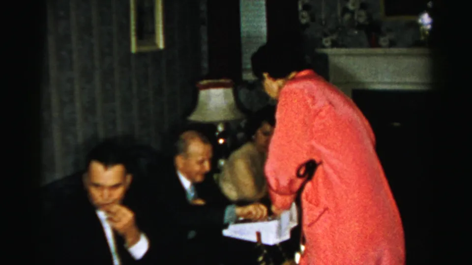 Albany 1950s living-room scene with suited men seated close together as a woman in red reaches toward a white box.