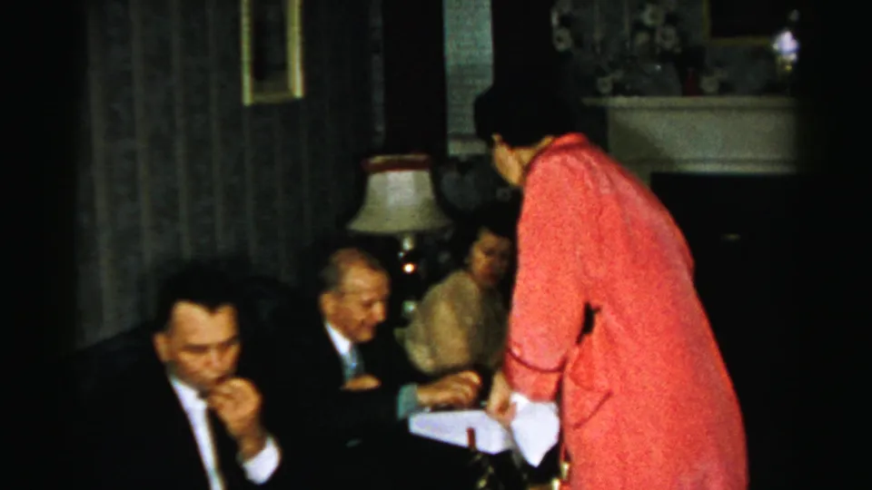 1950s Albany home-movie frame showing a woman in red holding a white box beside suited guests in a dim living room.