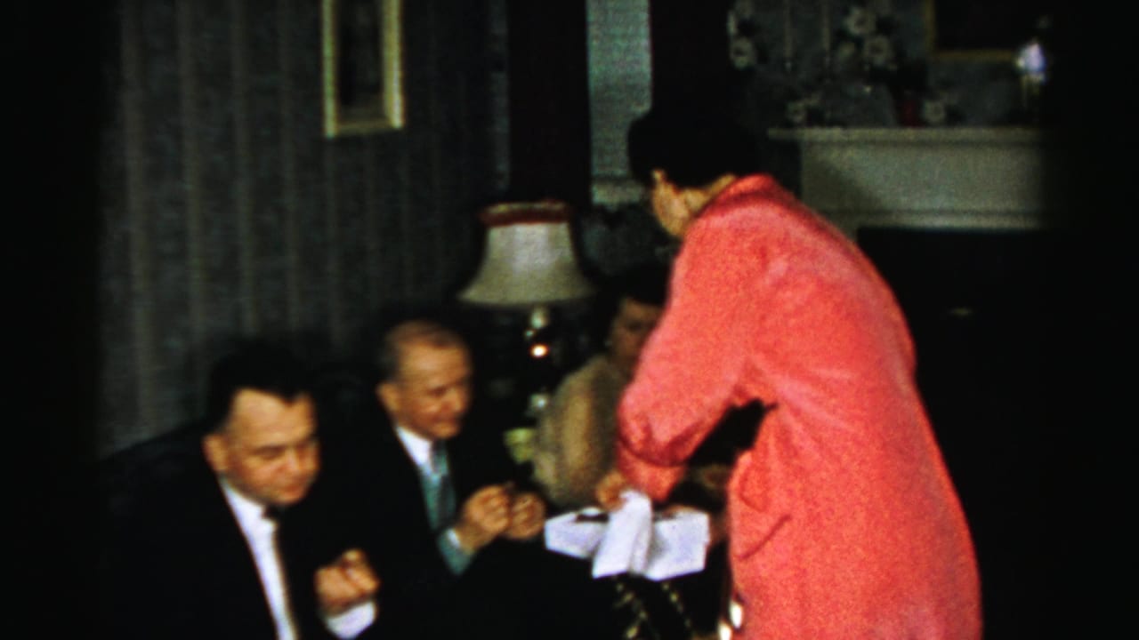Woman in a pink dress offers treats to seated adults in a dim sitting room.