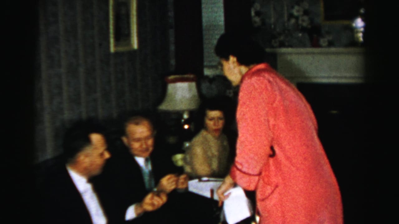 1956 Albany sitting-room gathering with suited men and women seated while a woman in pink offers refreshments from a white box.