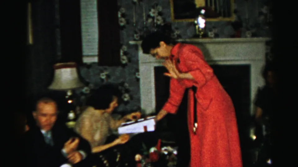 Home-movie frame from a 1956 Albany indoor gathering, with a woman serving treats beside seated guests in a decorated sitting room.