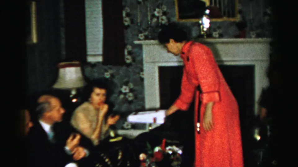 1956 Albany home-movie frame showing a woman standing in a pink dress near seated adults, with fireplace and floral wallpaper behind them.
