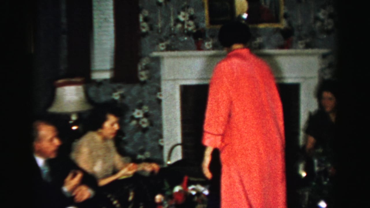 1956 Albany home-movie frame showing adults around a fireplace while a woman in a red robe stands with her back to the camera.