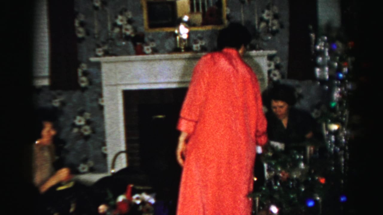 Woman in a red robe stands beside a tinsel Christmas tree and seated adults near the fireplace.