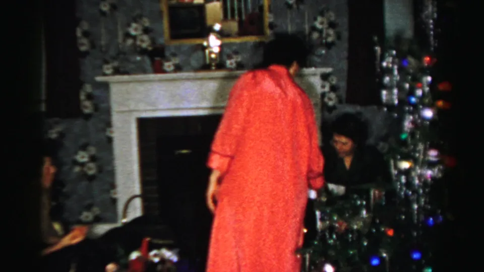 Home-movie frame from the Albany holiday gathering showing seated adults, a standing woman in red, and a decorated tinsel tree.