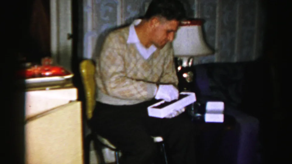 1956 Albany home-movie frame of a man in a patterned sweater seated in the living room with an opened box or gift in his hands.