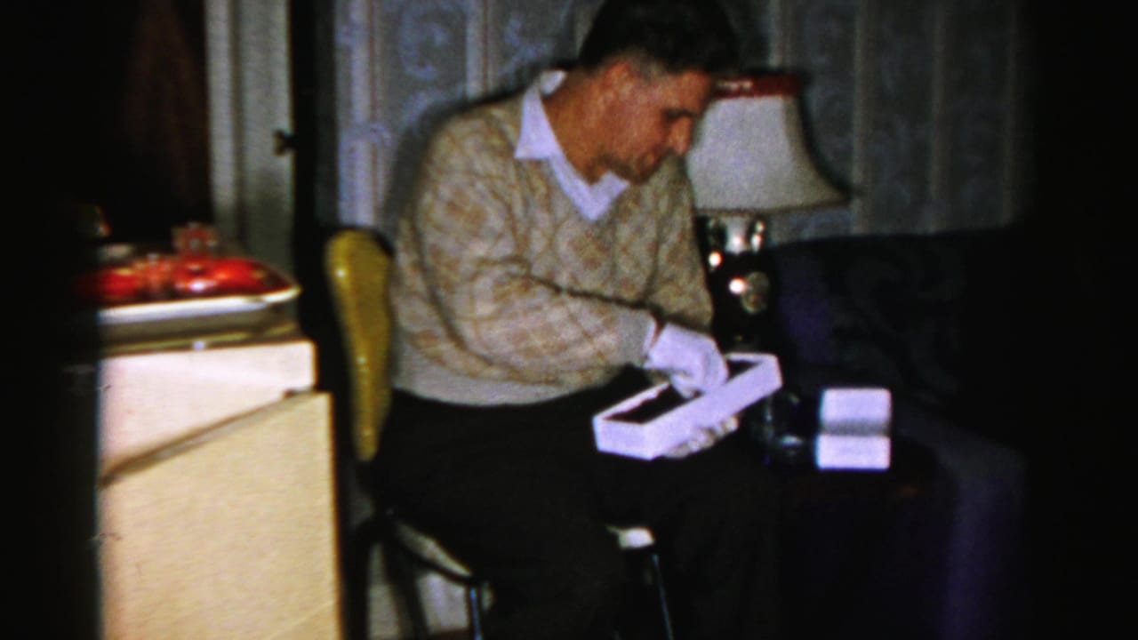 Man in a patterned sweater and white gloves sits with a white box in a dim room.