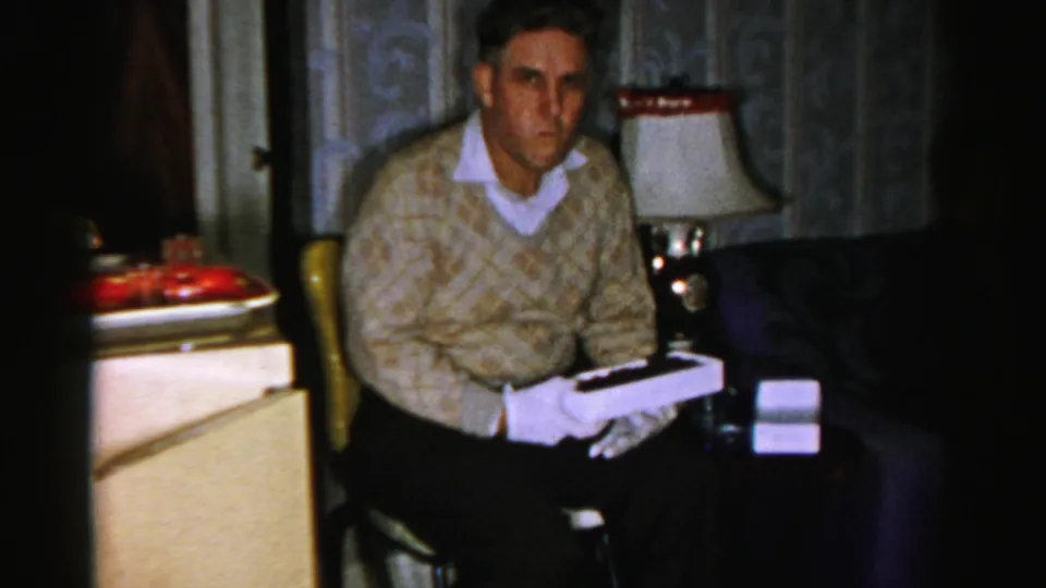 The man looks toward the camera from a chair, holding the open white box in a 1956 Albany domestic interior.