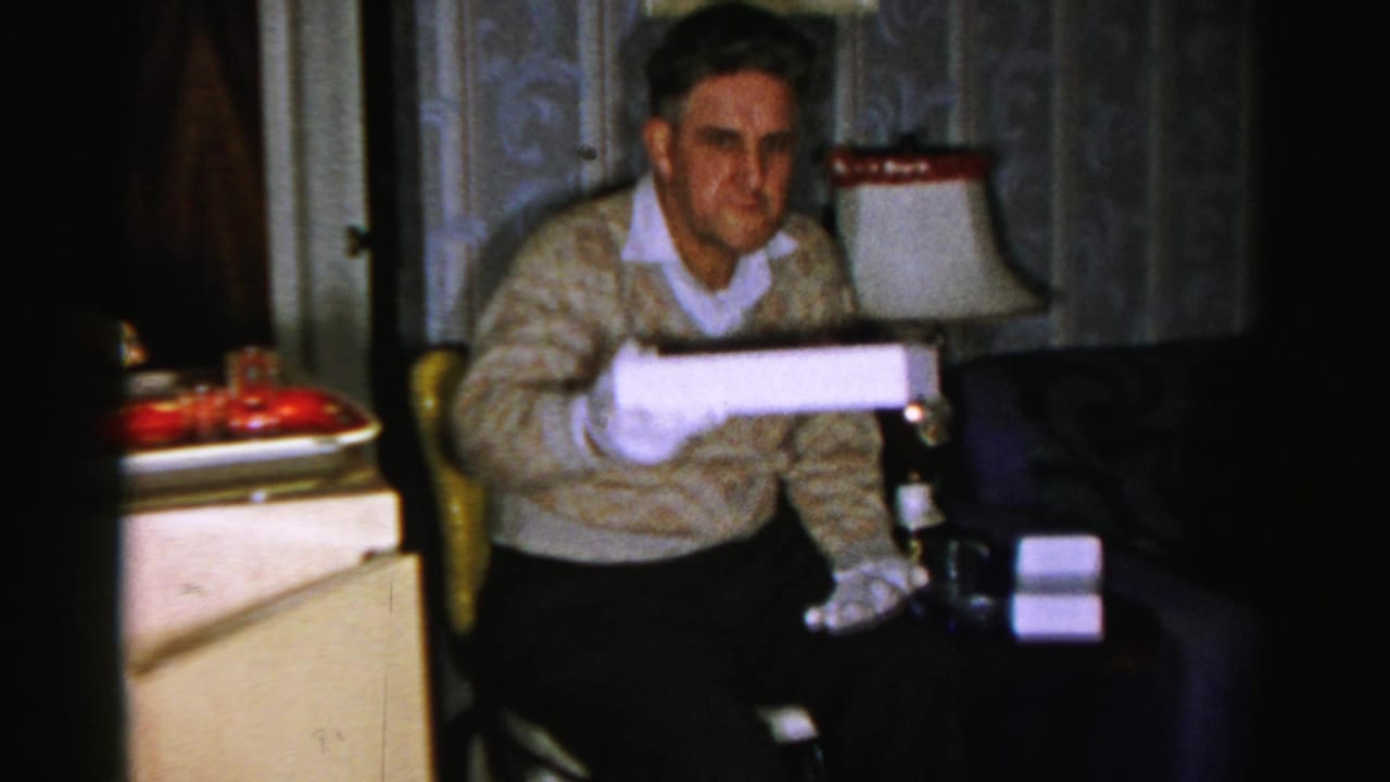 In an Albany home-movie frame from 1956, an older man in white gloves holds a white box beside a lamp and patterned wallpaper.