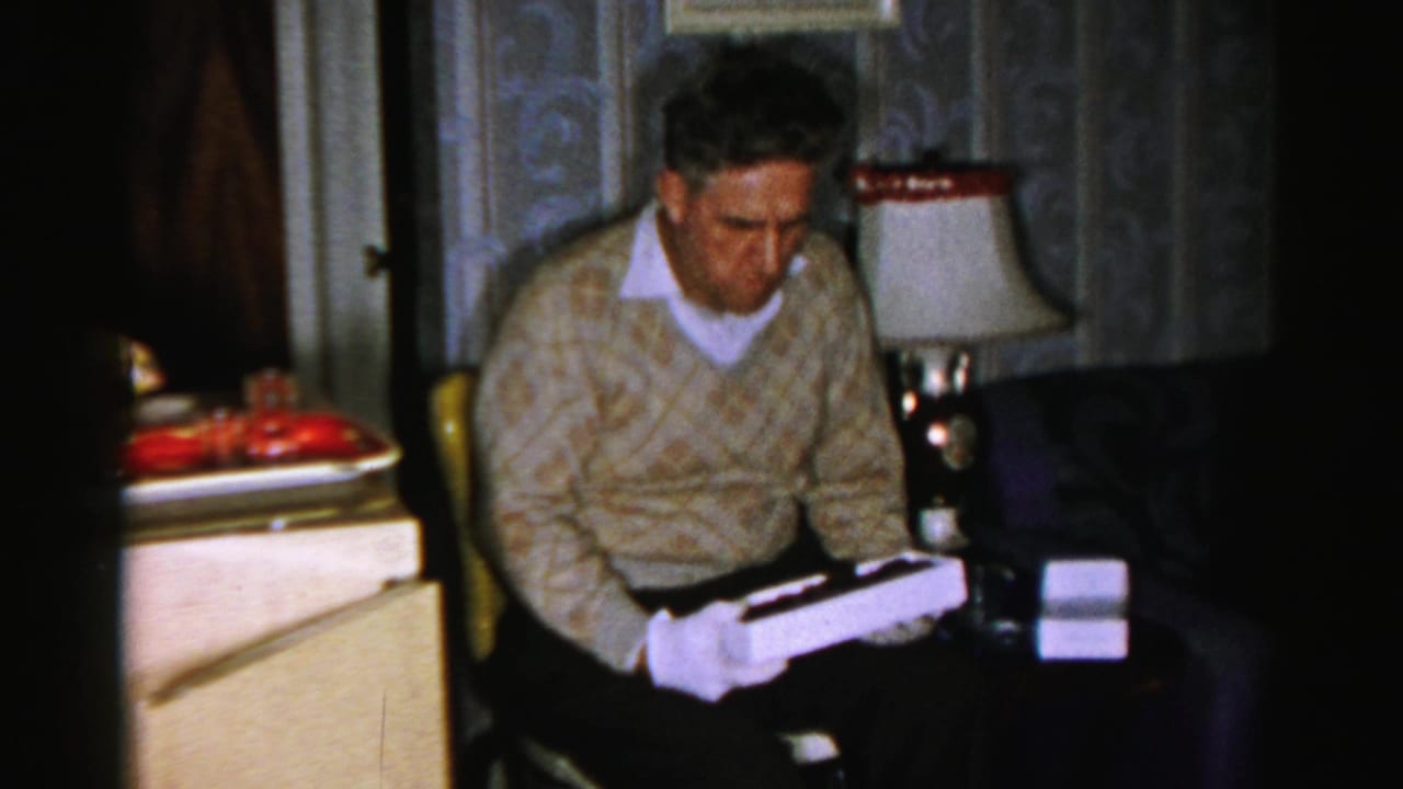 Older man looks down at a white box while seated near a lamp and side table.