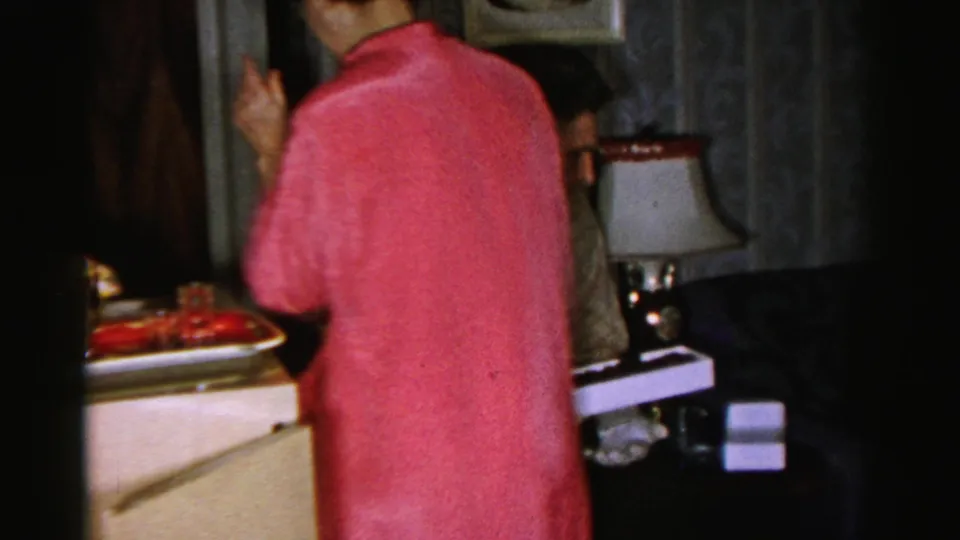 A woman in a pink coat briefly enters the 1956 Albany home-movie frame, partly blocking the seated man and the side table.