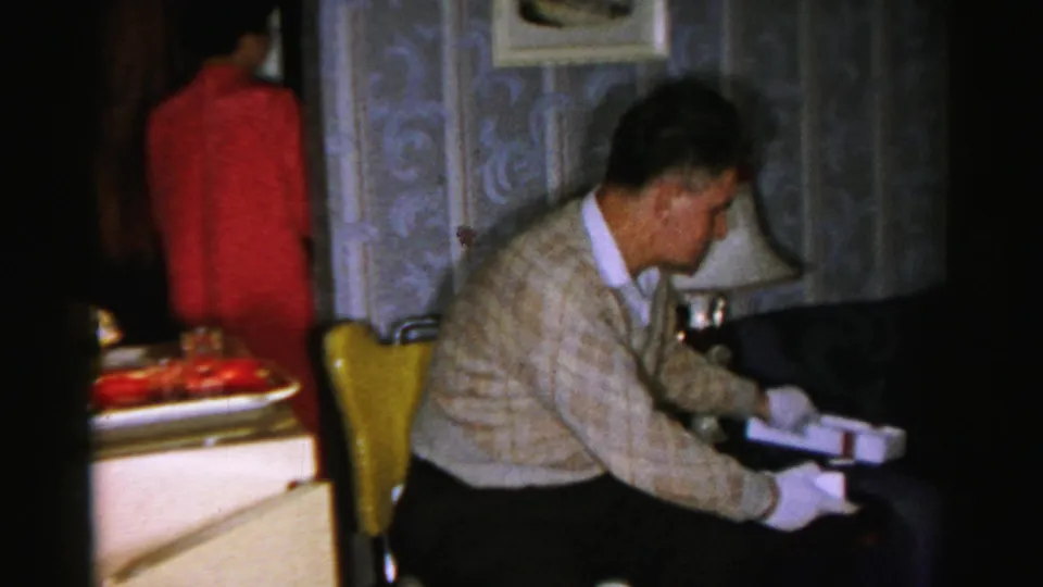 The older man turns to his right with the white box near a chair, while the woman in pink is visible in the background.