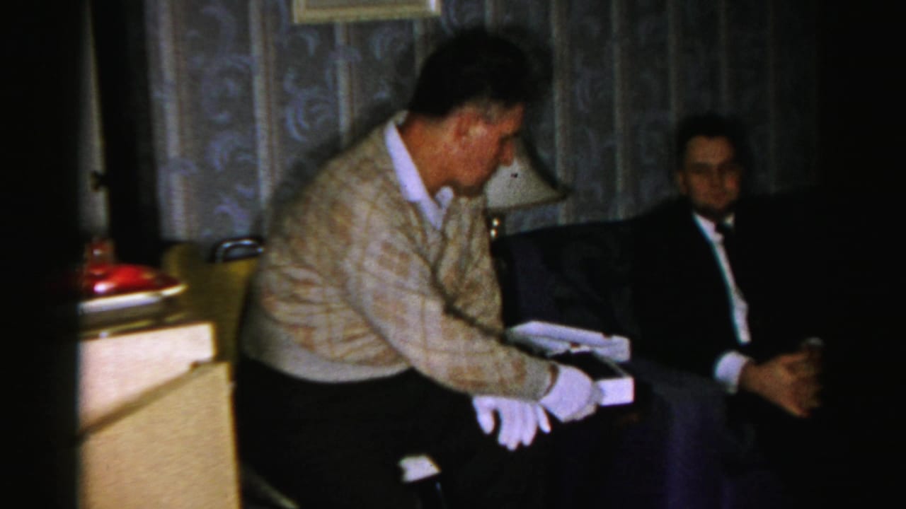 Two men in a dim living room sit beside a lamp as one handles a white candy box.