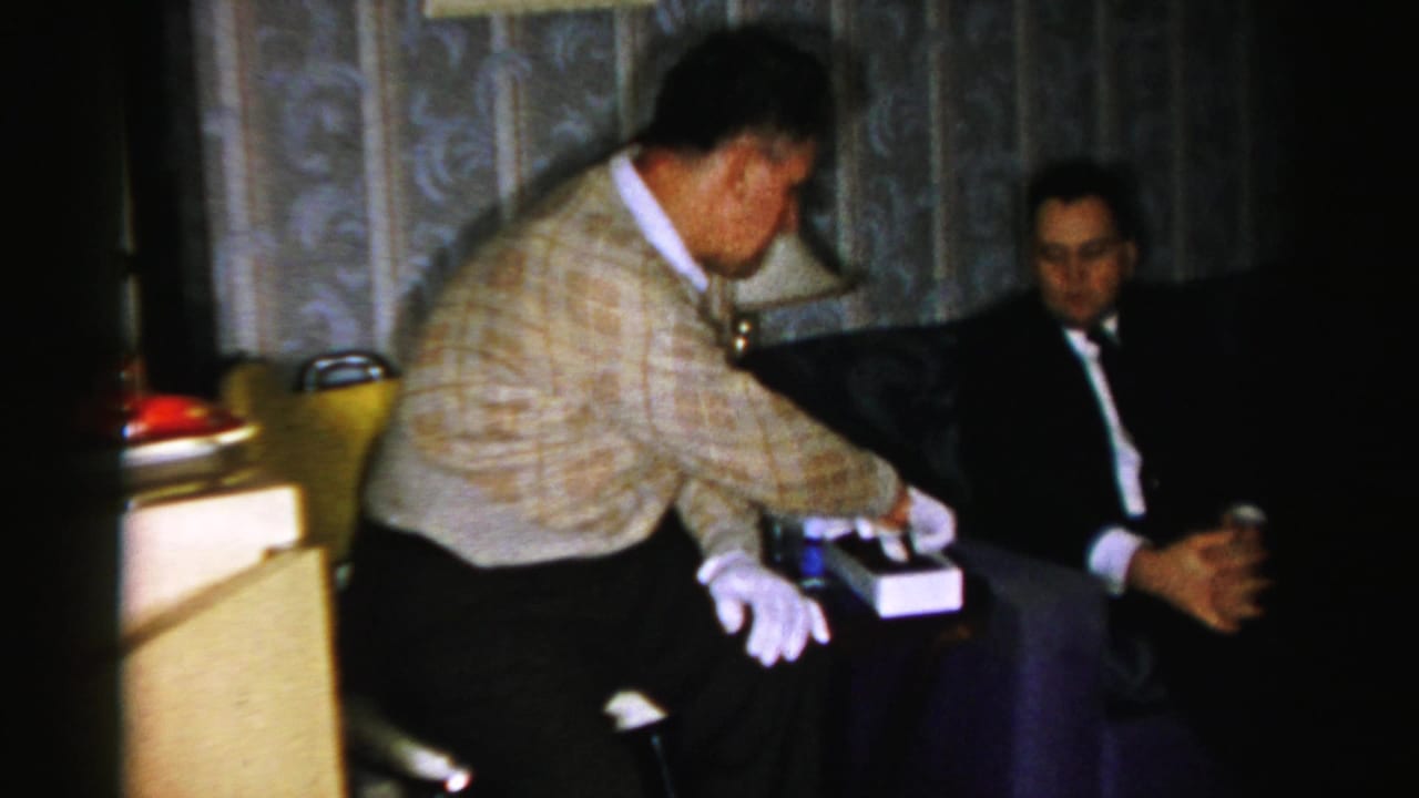 A standing man reaches toward a candy box while another man sits in a dark suit.
