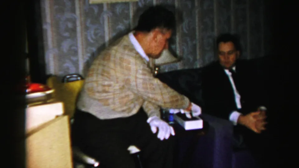 The man in the patterned sweater leans over the candy box while the suited man watches in a dim Albany living room.