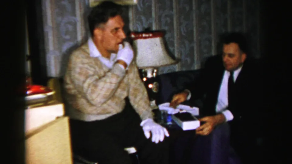 A seated man holds the white candy box as the man in the patterned sweater raises a hand near his mouth in the 1956 home-movie scene.