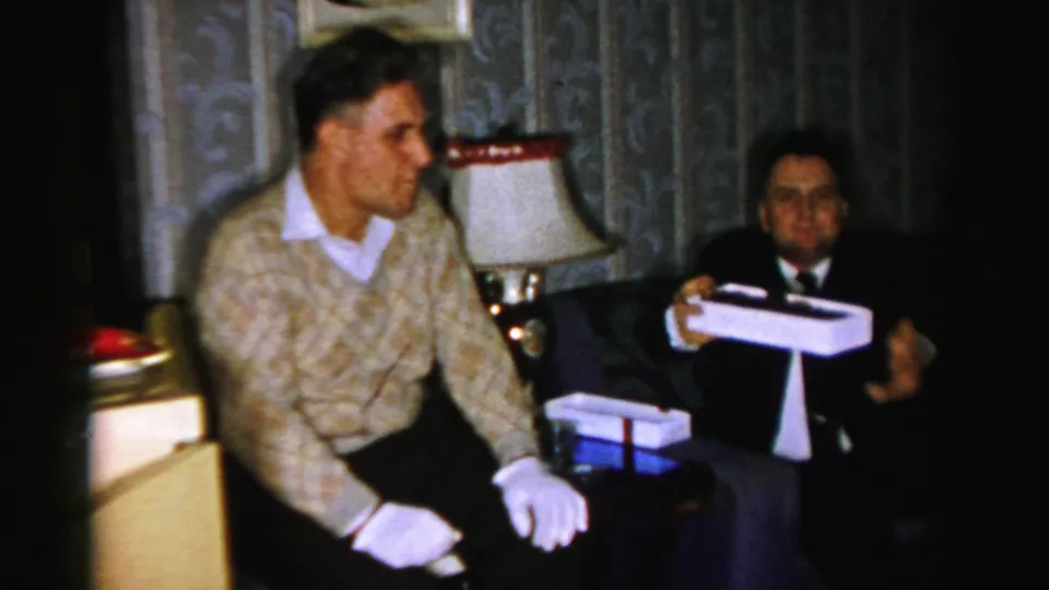 The suited man holds up the white candy box toward the camera or companion while the man in the patterned sweater sits beside him.