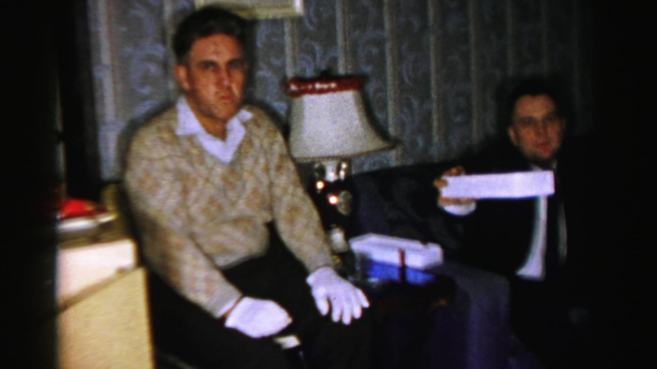 Two men seated indoors, one in a patterned sweater and white gloves, with curtains and a lamp behind them.