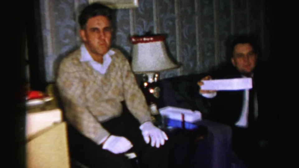 Albany home-movie frame from the 1950s showing two seated men in a dim private room, one wearing white gloves and a patterned sweater.