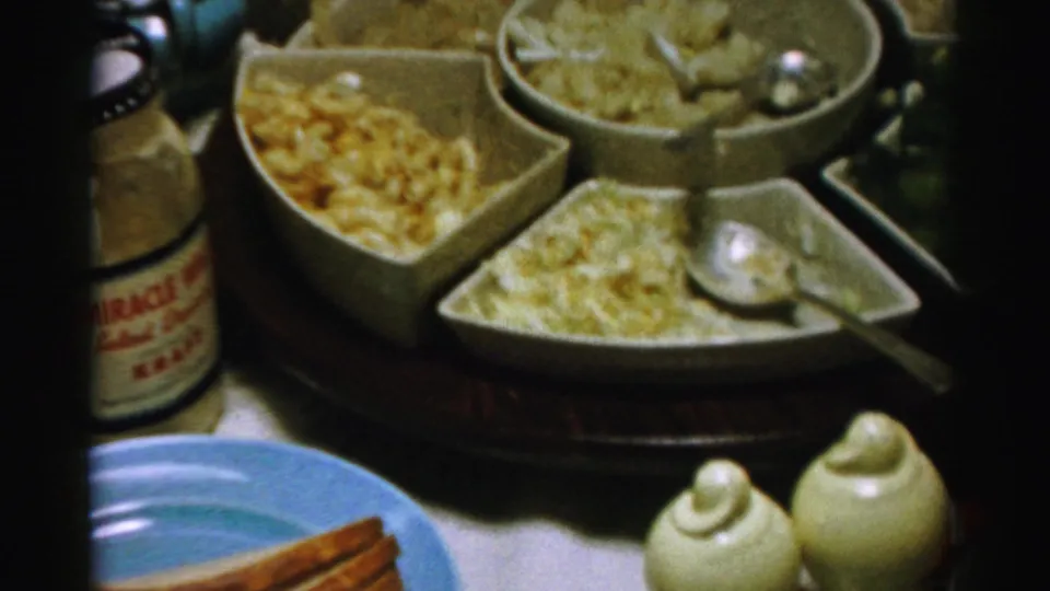Close 1950s Albany home-movie view of appetizer dishes, ceramic shakers, crackers, and a partially visible labeled bottle on a private table.