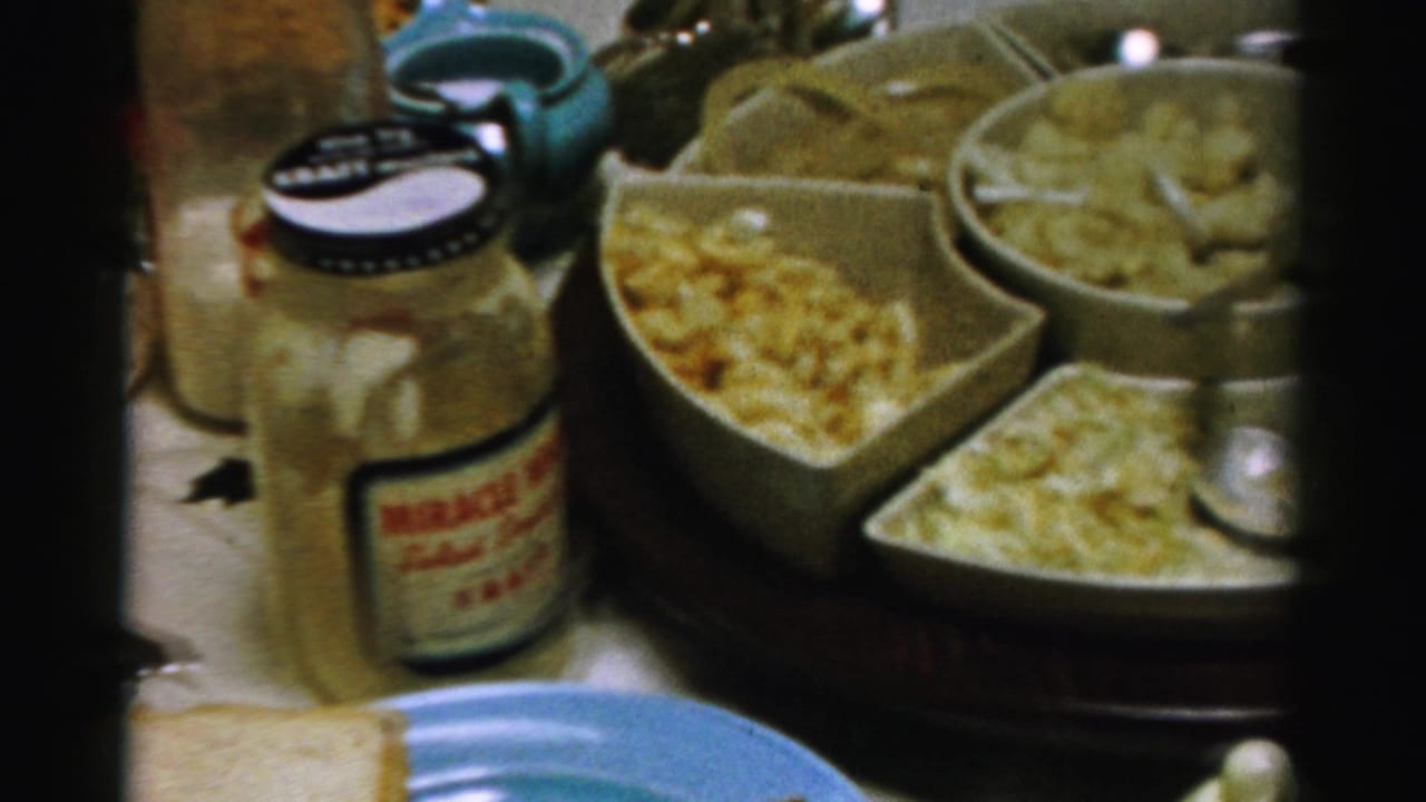 1956 Albany home-movie frame showing a holiday table with a Miracle Whip jar, macaroni salad, bread, and pastel blue dishes.