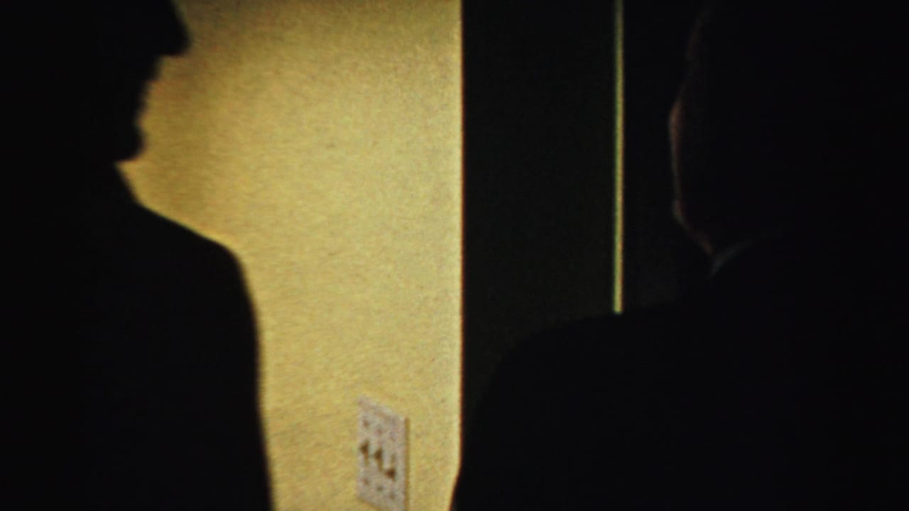 A man in a hat is seen in dark profile beside a wall and switch plate.