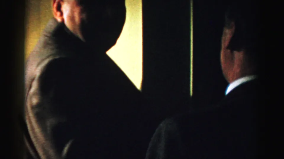 Two adult men in dark suits stand in a low-light Albany room, with one partly facing the camera and another seen from behind.