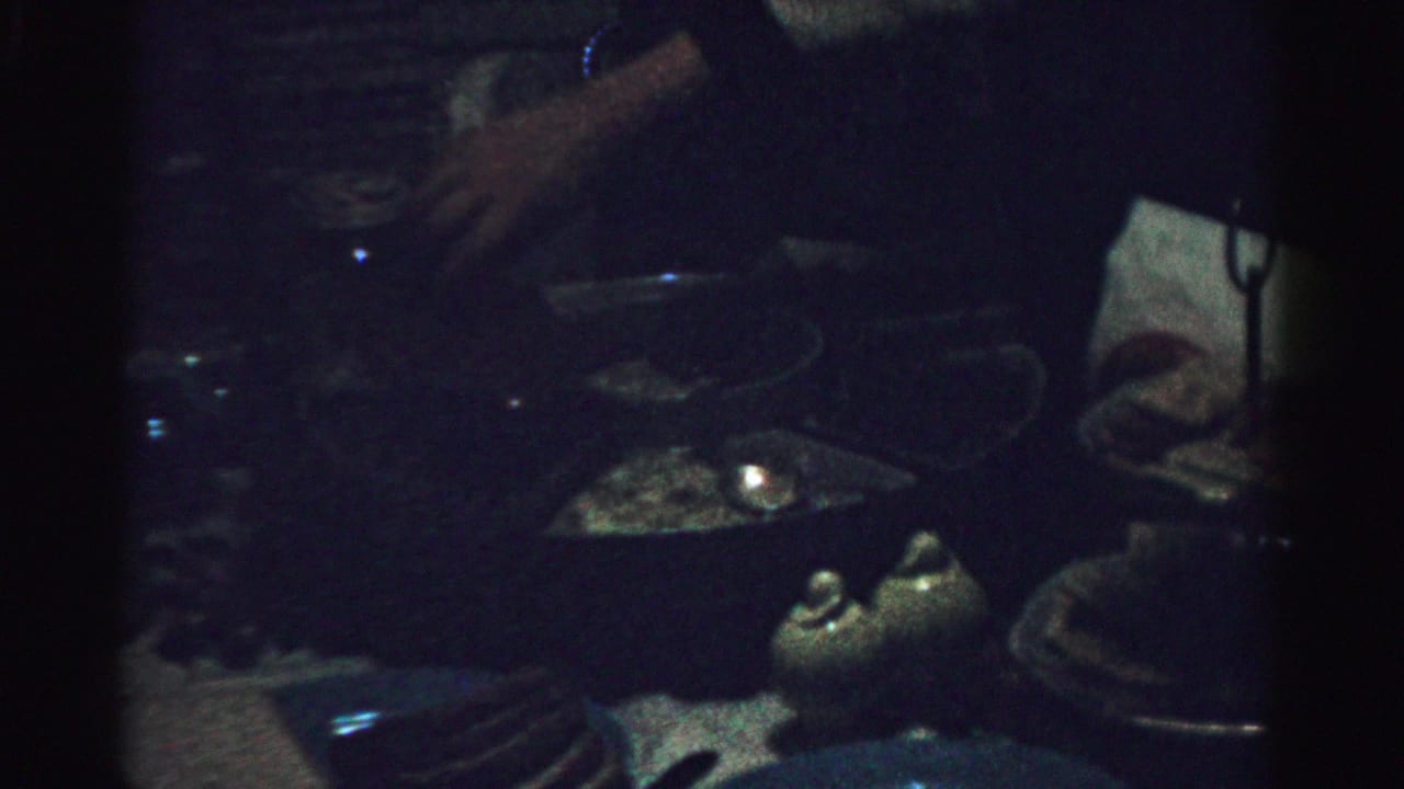 Albany-area 1950s home-movie frame showing hands near plates, glassware, and a candlelit table in a dark private interior.