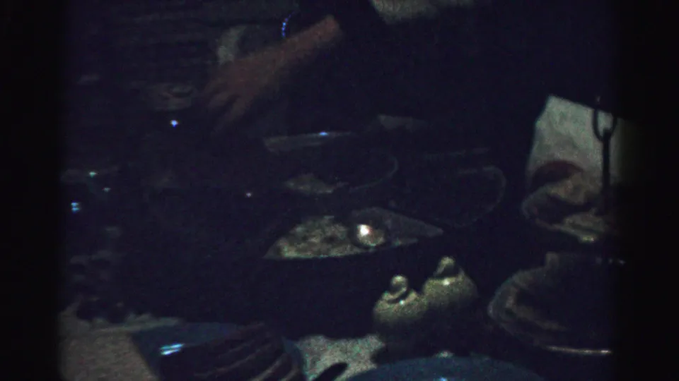 Albany-area 1950s home-movie frame showing hands near plates, glassware, and a candlelit table in a dark private interior.