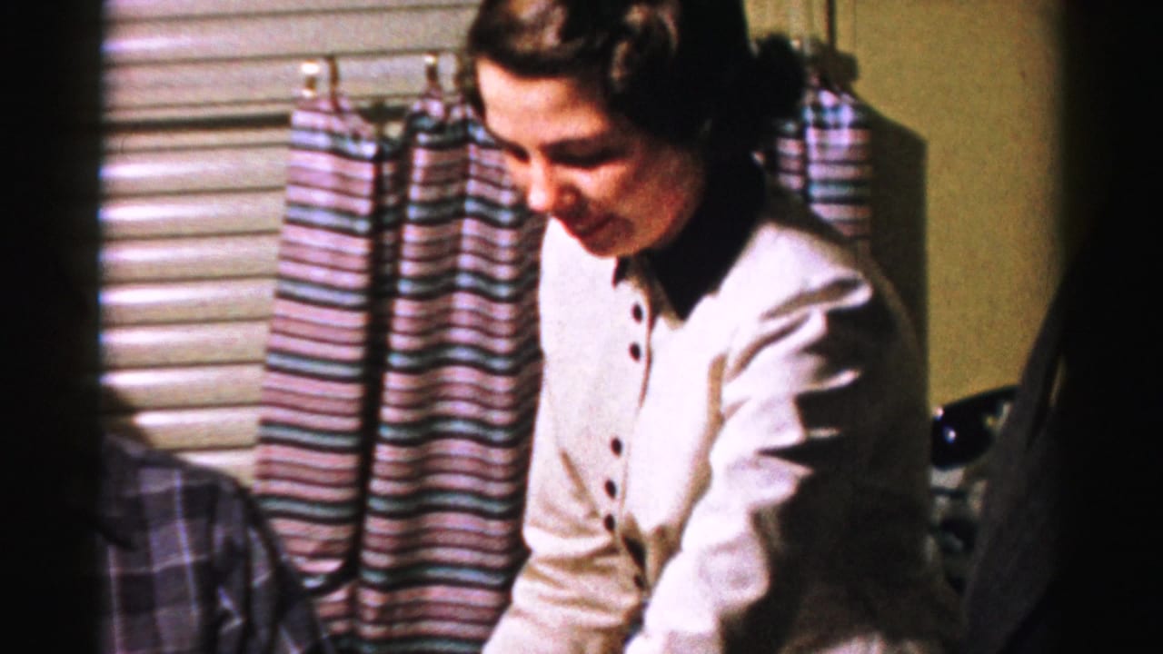 In Albany home-movie footage from the 1950s, a woman in white stands near striped curtains and window blinds during a private gathering.