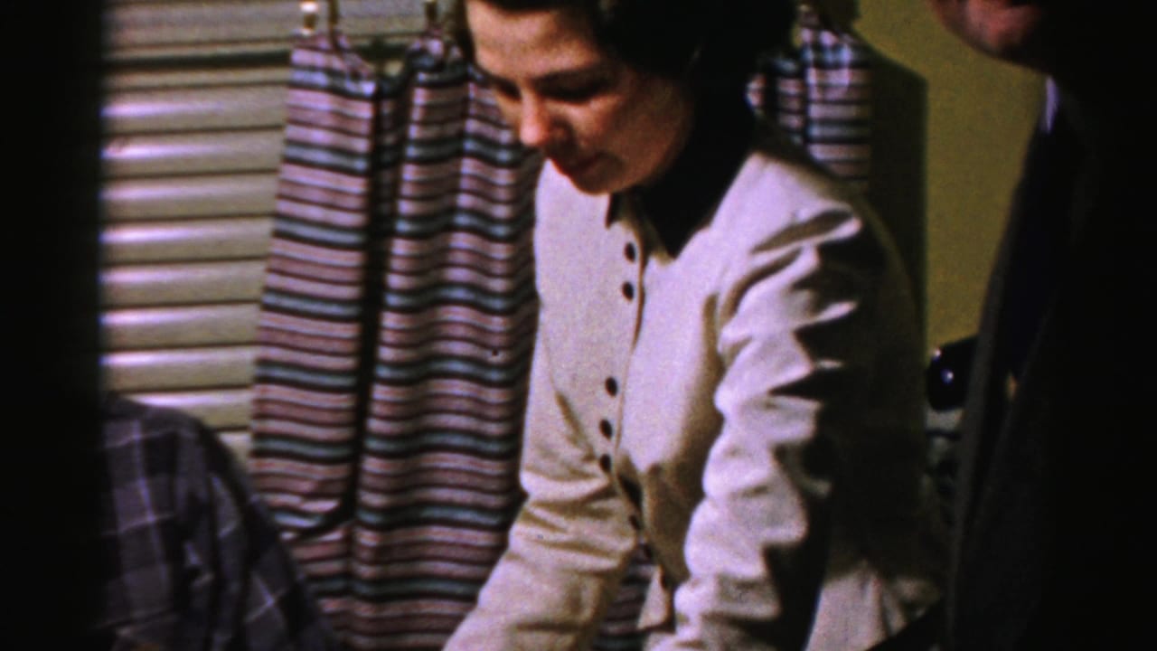 Woman looks downward near curtains while a suited guest stands beside her.