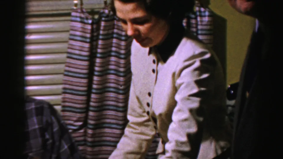 The woman in white looks down in a 1950s Albany interior as a suited guest appears at the edge of the home-movie frame.