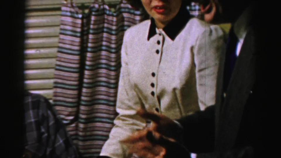 A woman in white faces the camera area while a guest's hand gestures in the foreground of the 1950s Albany home gathering.
