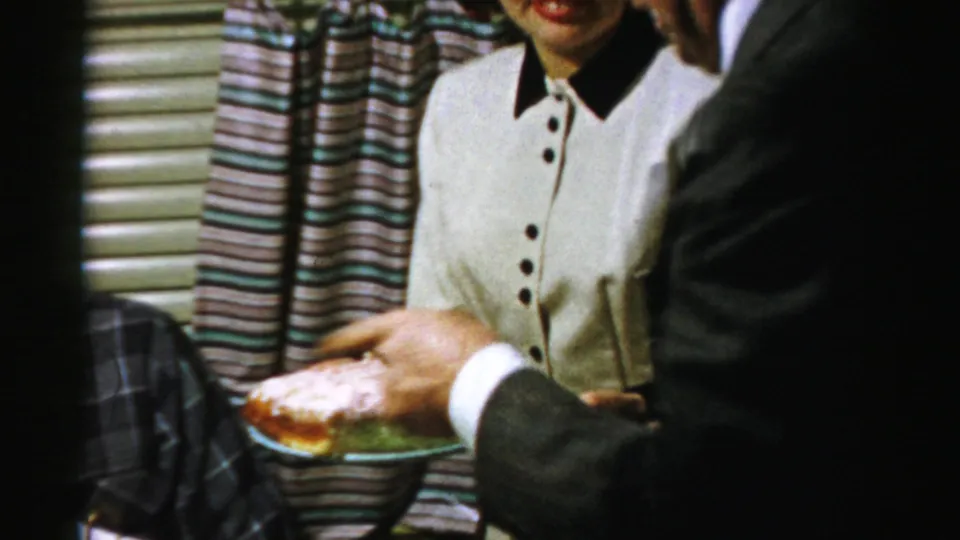 A suited guest holds a plate with pie near the woman in white during a 1950s Albany private home gathering.