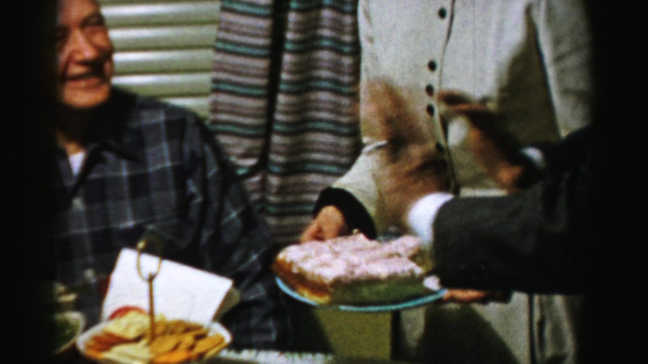 In this 1956 Albany home-movie frame, adults stand around a food table while a seated man in plaid smiles beside a pink-frosted cake.