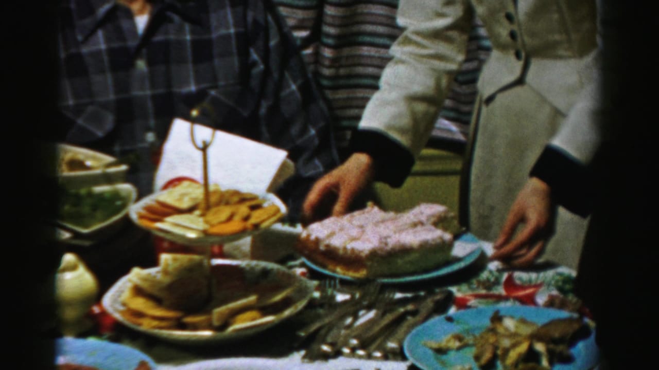Close view of a crowded table with cake, snack tray, plates, utensils, and adults nearby.