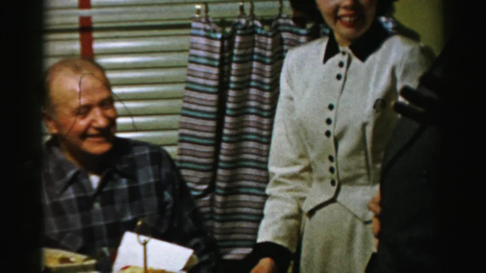 A seated older man in a plaid shirt smiles while a woman in a light dress stands near the table in a private Albany interior.