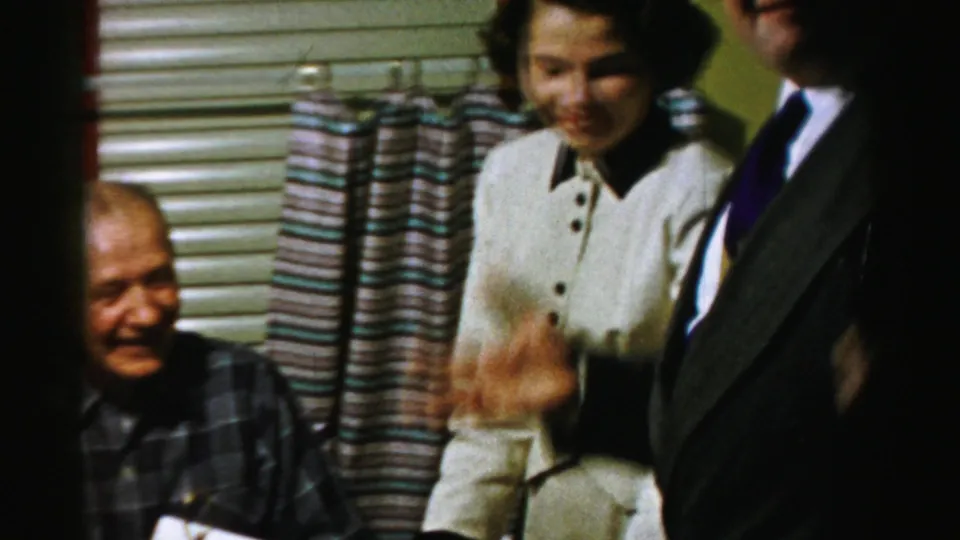 Adults in dress clothing smile and gesture beside the food table in this 1956 Albany home-movie party scene.