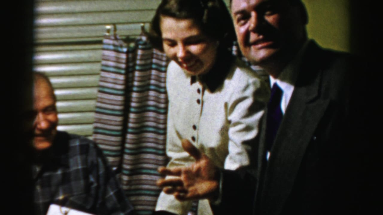 In a 1956 Albany home-movie frame, a young woman in a white dress and a suited man laugh beside an older man at a food table.
