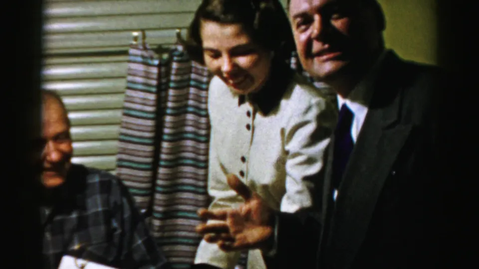 In a 1956 Albany home-movie frame, a young woman in a white dress and a suited man laugh beside an older man at a food table.
