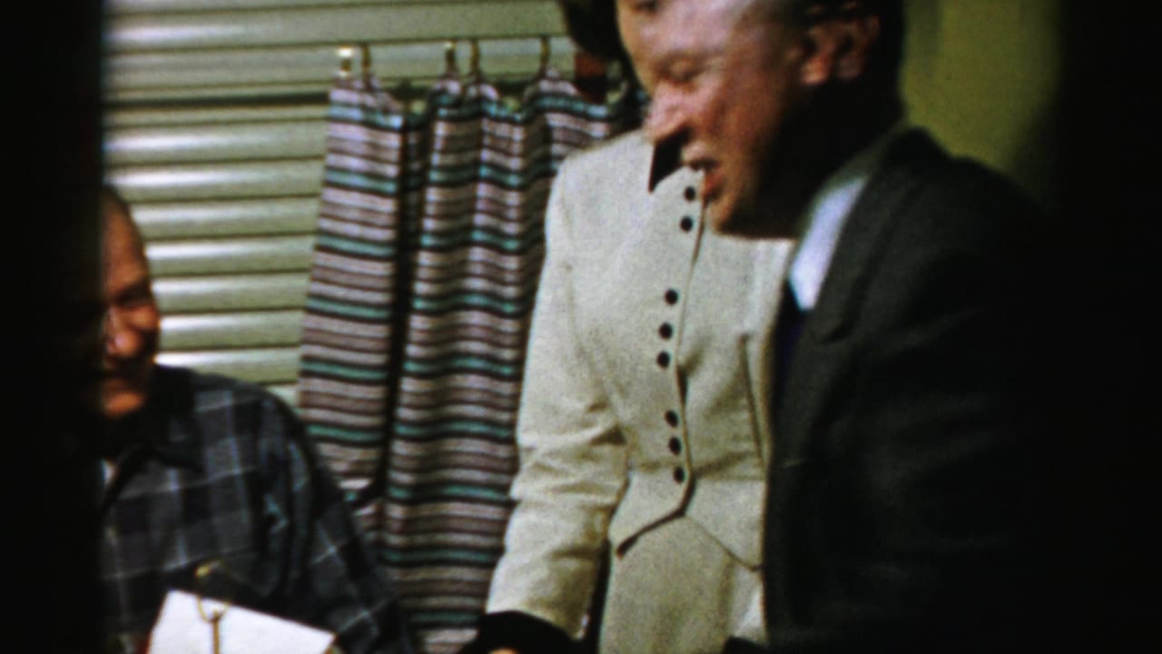 A man in a suit leans toward a woman in a white dress beside a seated older man.