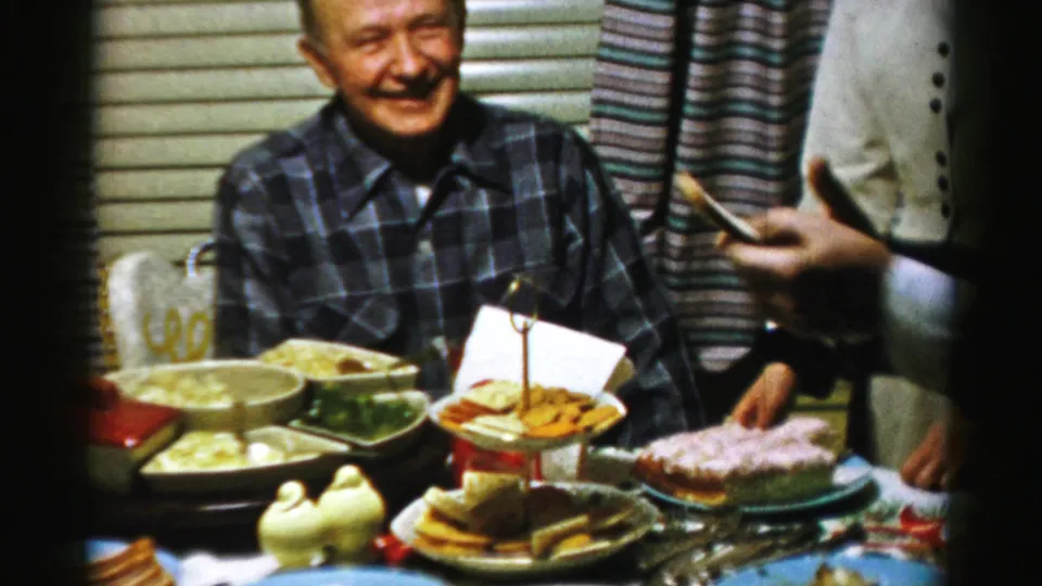 An older man in a plaid shirt smiles at a 1956 Albany indoor gathering, with dishes, platters, and cake-like food on the table.
