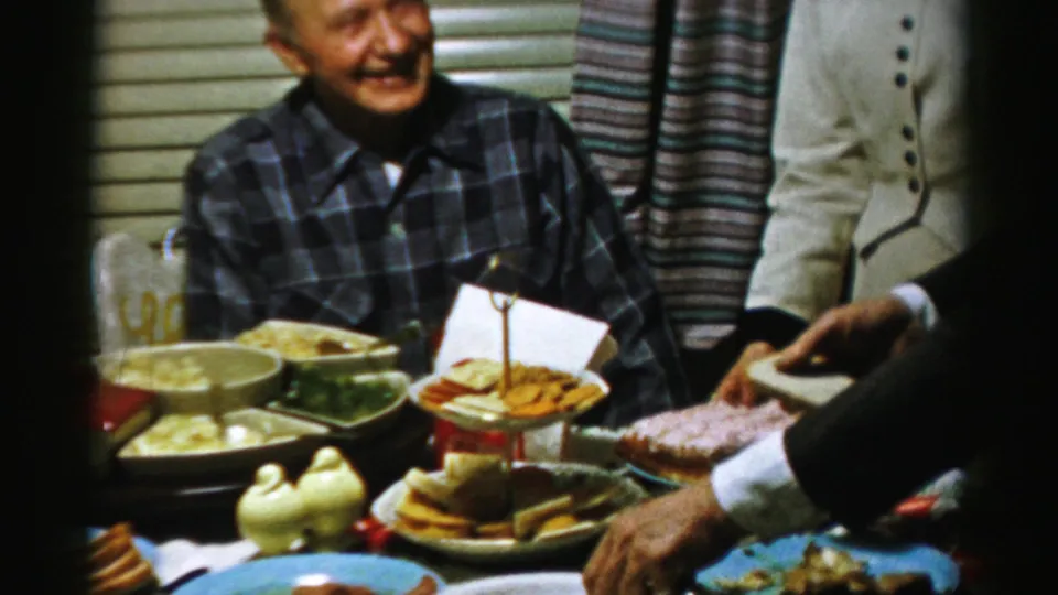 The older man smiles at the Albany family table as another adult reaches toward the dishes and a woman in white stands nearby.