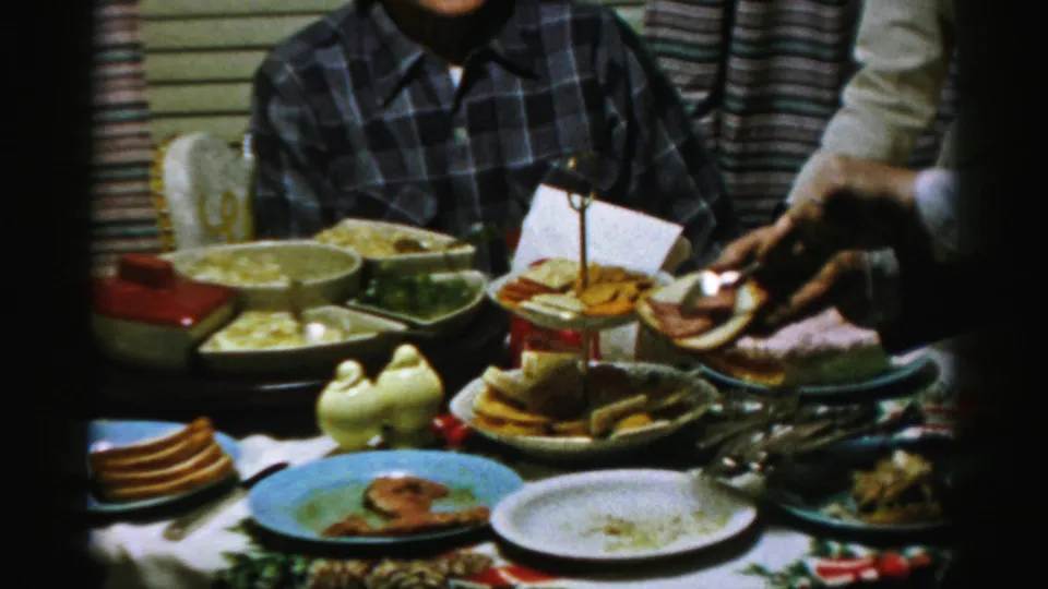 1950s Albany home-movie frame showing a hand holding bread above a crowded table with crackers, plates, serving dishes, and a seated adult nearby.