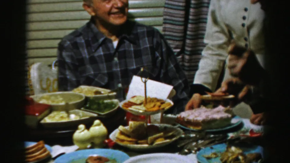 1950s Albany family dinner frame with a smiling seated man, a standing adult, and hands preparing bread beside dishes and serving utensils.