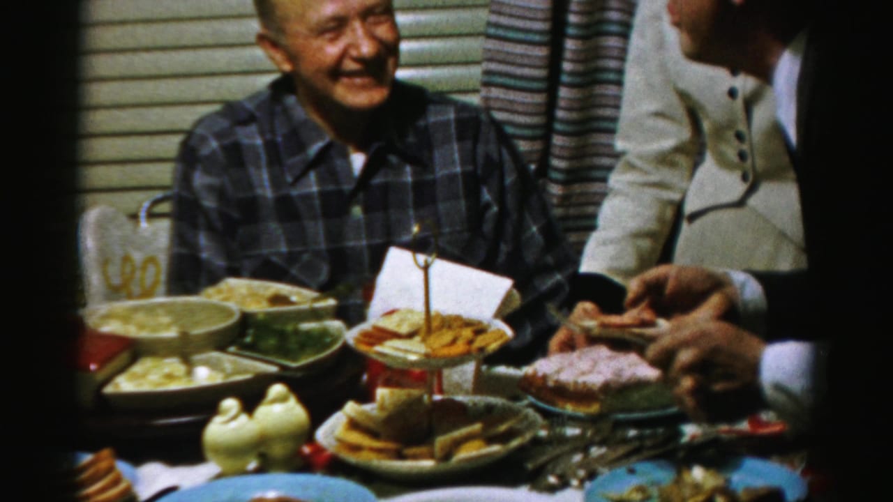 Adults seated around a table with serving dishes and dessert while a smiling man in a plaid shirt faces the camera.