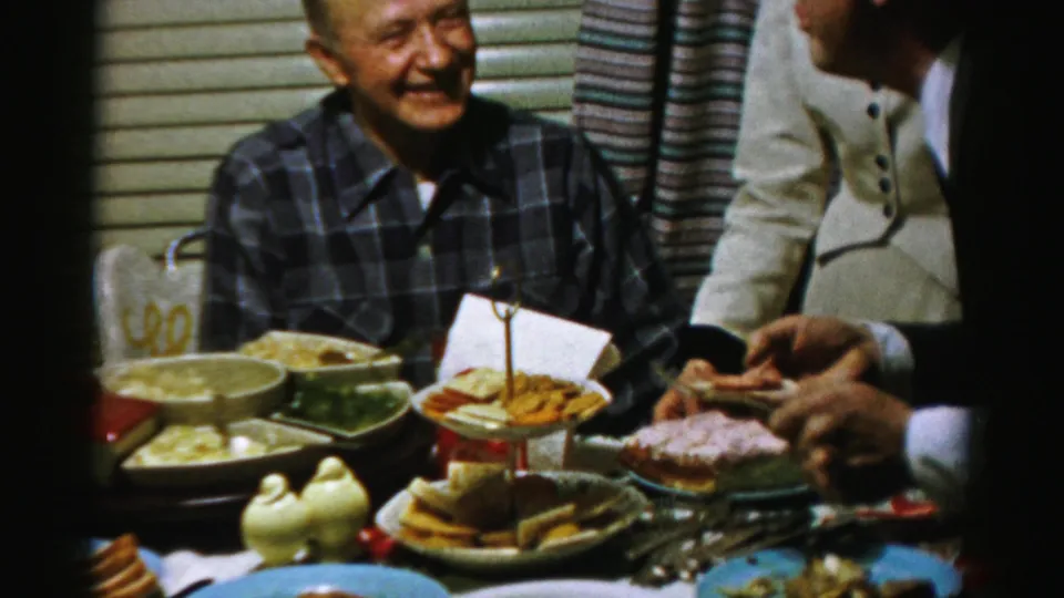 In an Albany 1950s home-movie frame, adults gather around a crowded table as a smiling man in a plaid shirt sits near dishes and dessert.